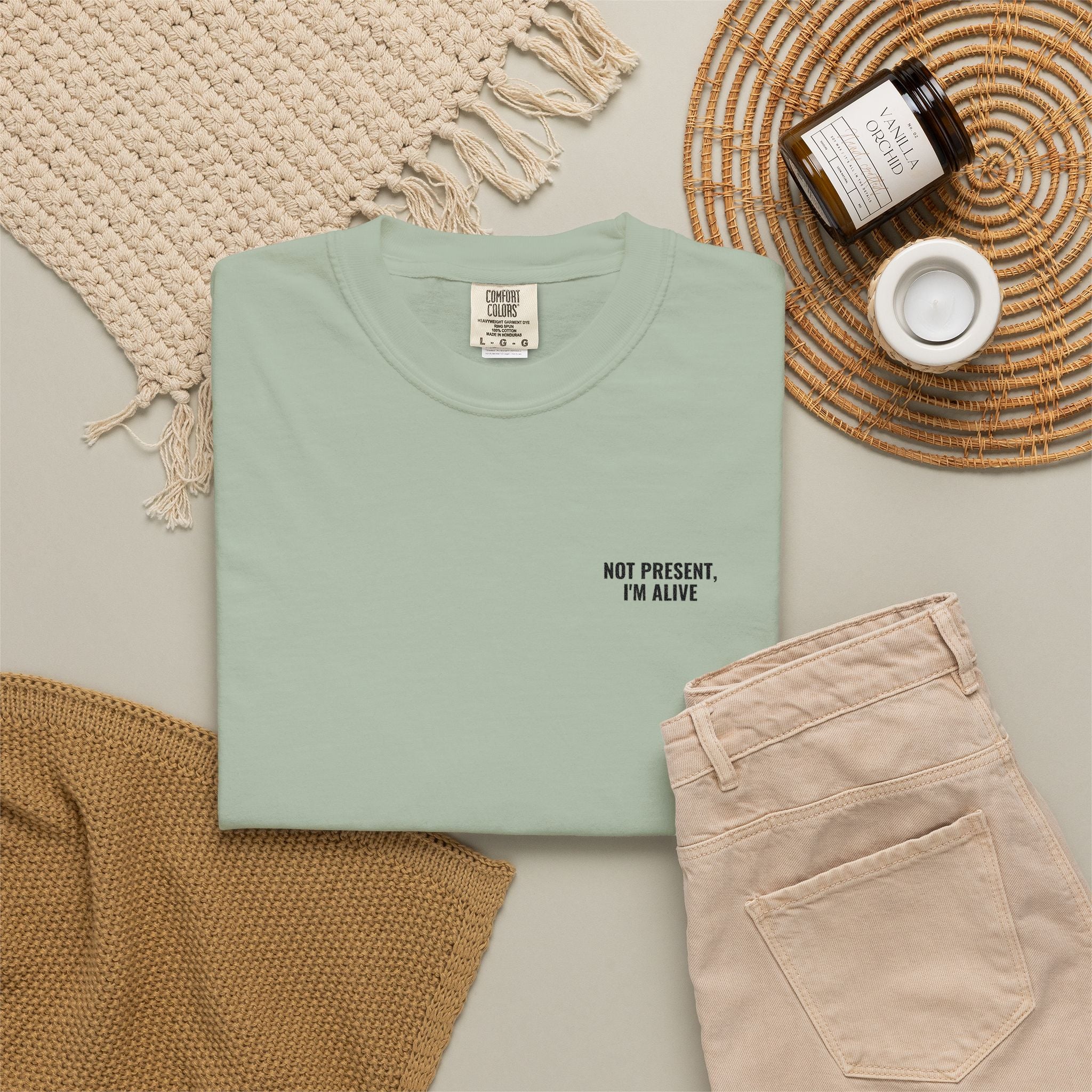 Folded light green t-shirt with text, beige pants, and decorative items on a neutral background