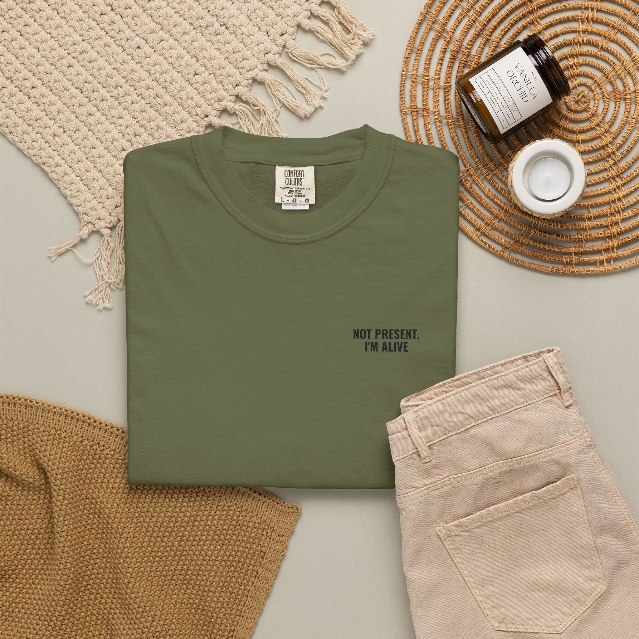 Green t-shirt with 'Not present, I'm alive' text on a neutral background with pants and decor items.