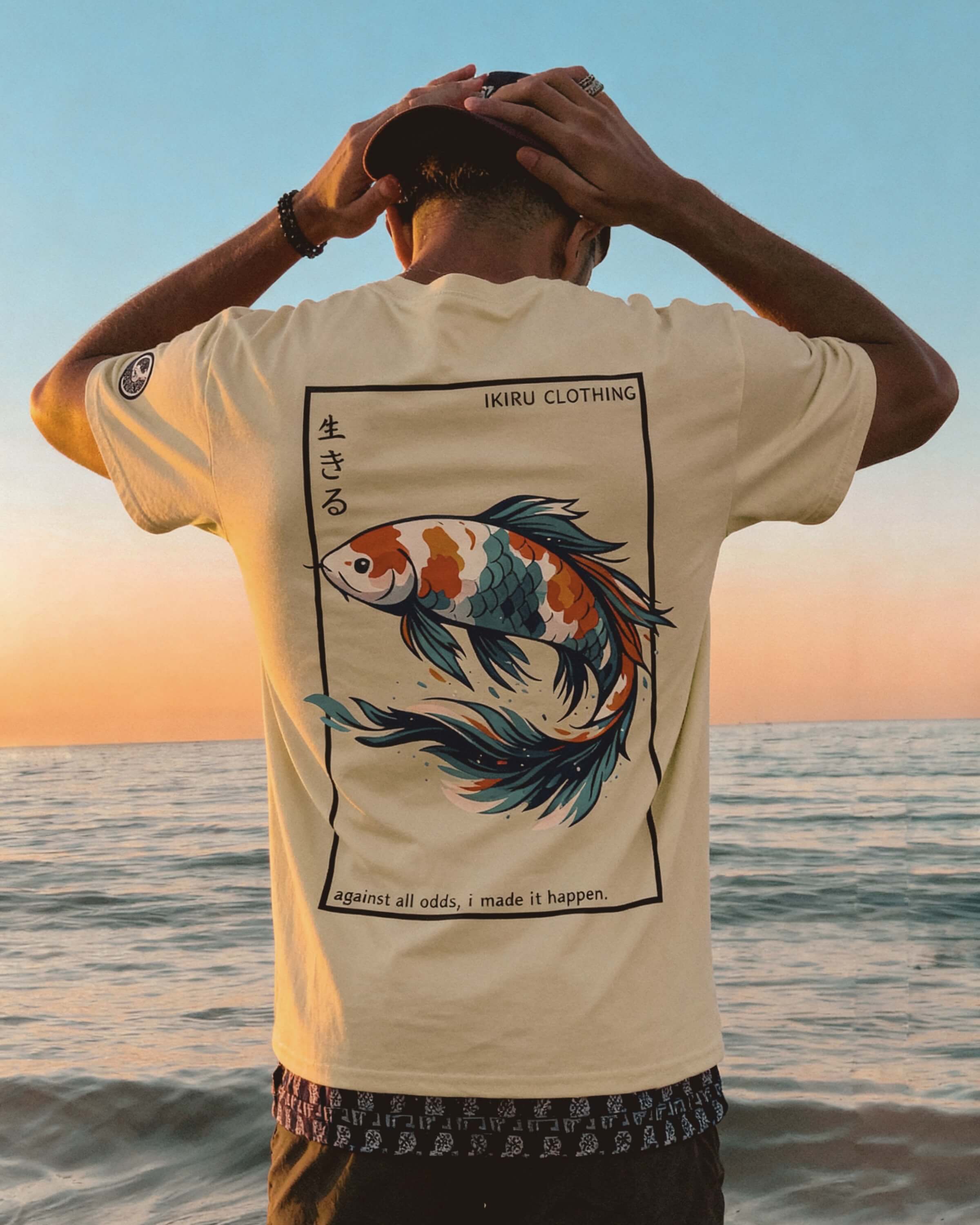 Person on a beach with amazing sunset wearing a beige shirt with IKIRU backprint design with his hands on his hands