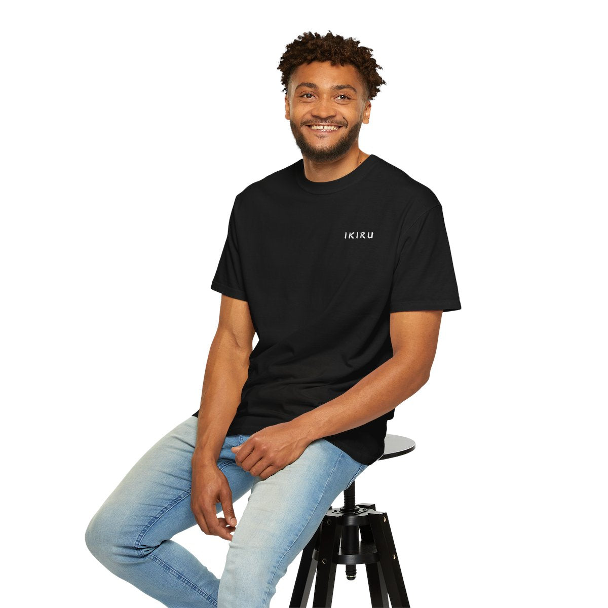 Man sitting on a stool wearing a black t-shirt with a logo and light blue jeans on a white background